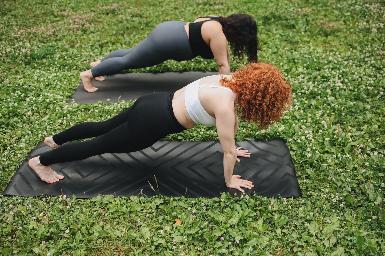 Two women holding a plank position on yoga mats outdoors in a lush green setting.